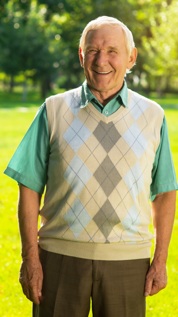 an elderly man in a garden wearing a jumper and smiling at the camera