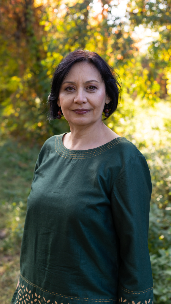 a photo of a middle-aged woman standing in a woods with trees behind her