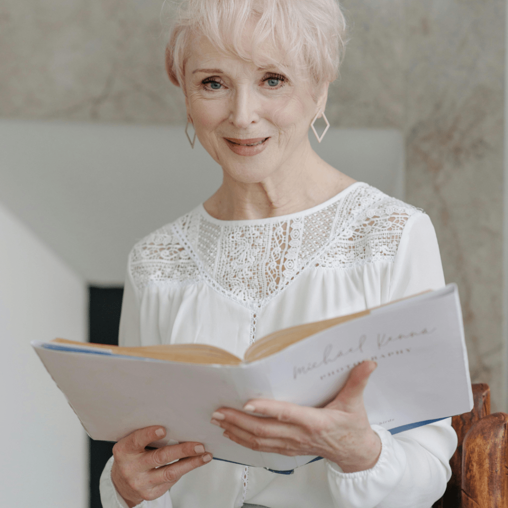 A PHOTO OF AN OLDER LADT WITH WHITE HAIR. SHE IS HOLDING A LARGE BOOK AND STARING AT THE CAMERA
